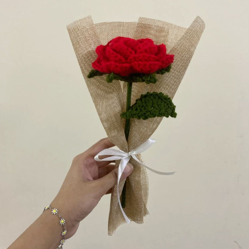 Hand holding a bouquet of red flowers wrapped in brown paper with a white ribbon against a plain background